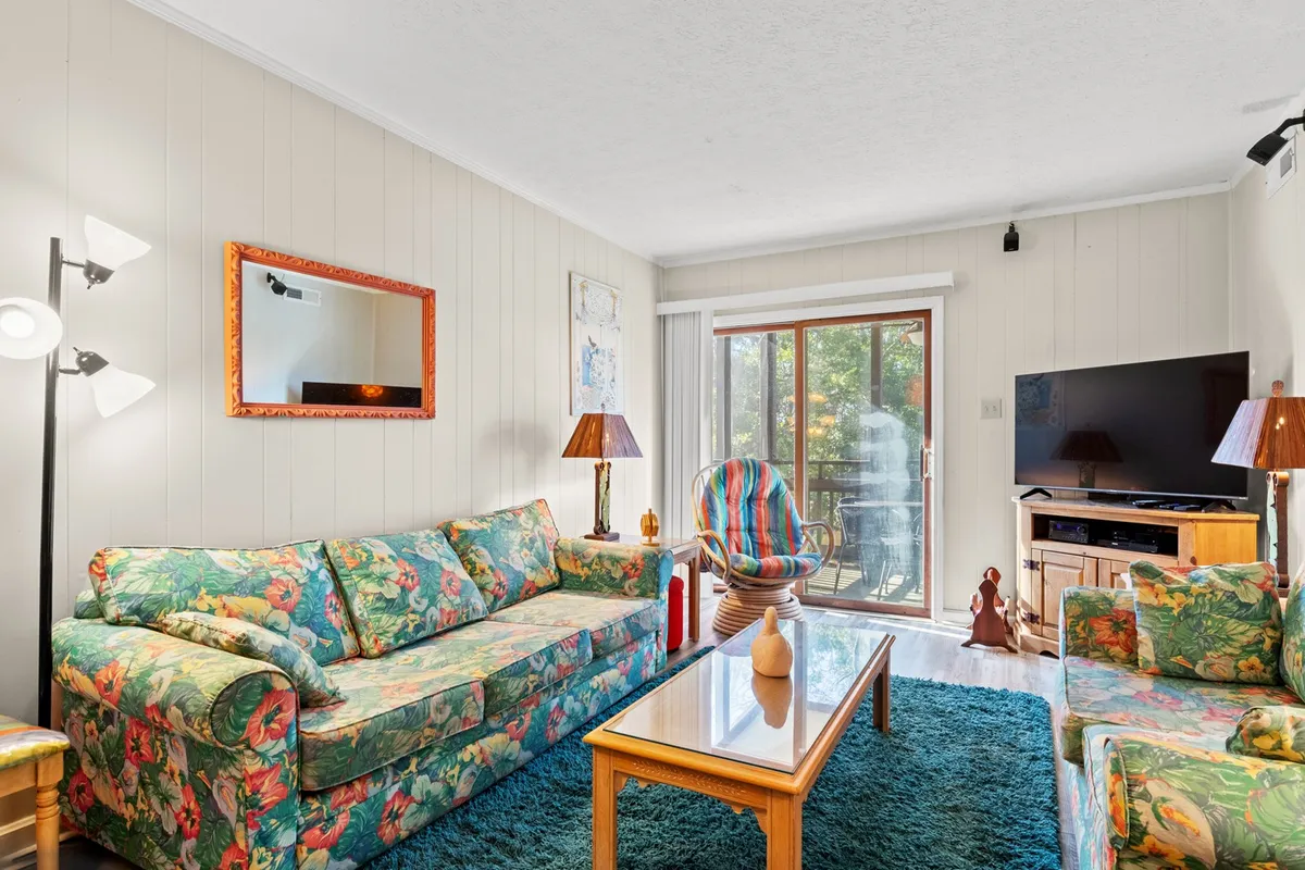 A bright and airy living room featuring floral-patterned sofas, a wooden coffee table, and a large window that lets in abundant natural light. A colorful accent chair adds vibrancy to the room, and the overall atmosphere is both cheerful and inviting.