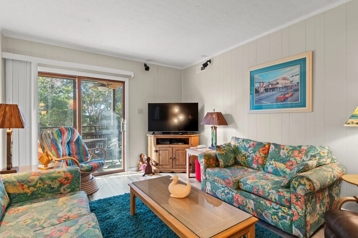 The image displays a bright, cheerful living room with colorful floral sofas, a wooden coffee table, tropical decor accents, and a large television. Sunlight pours in through a sliding glass door, creating a welcoming, relaxing space perfect for unwinding after a day at the nearby beach.