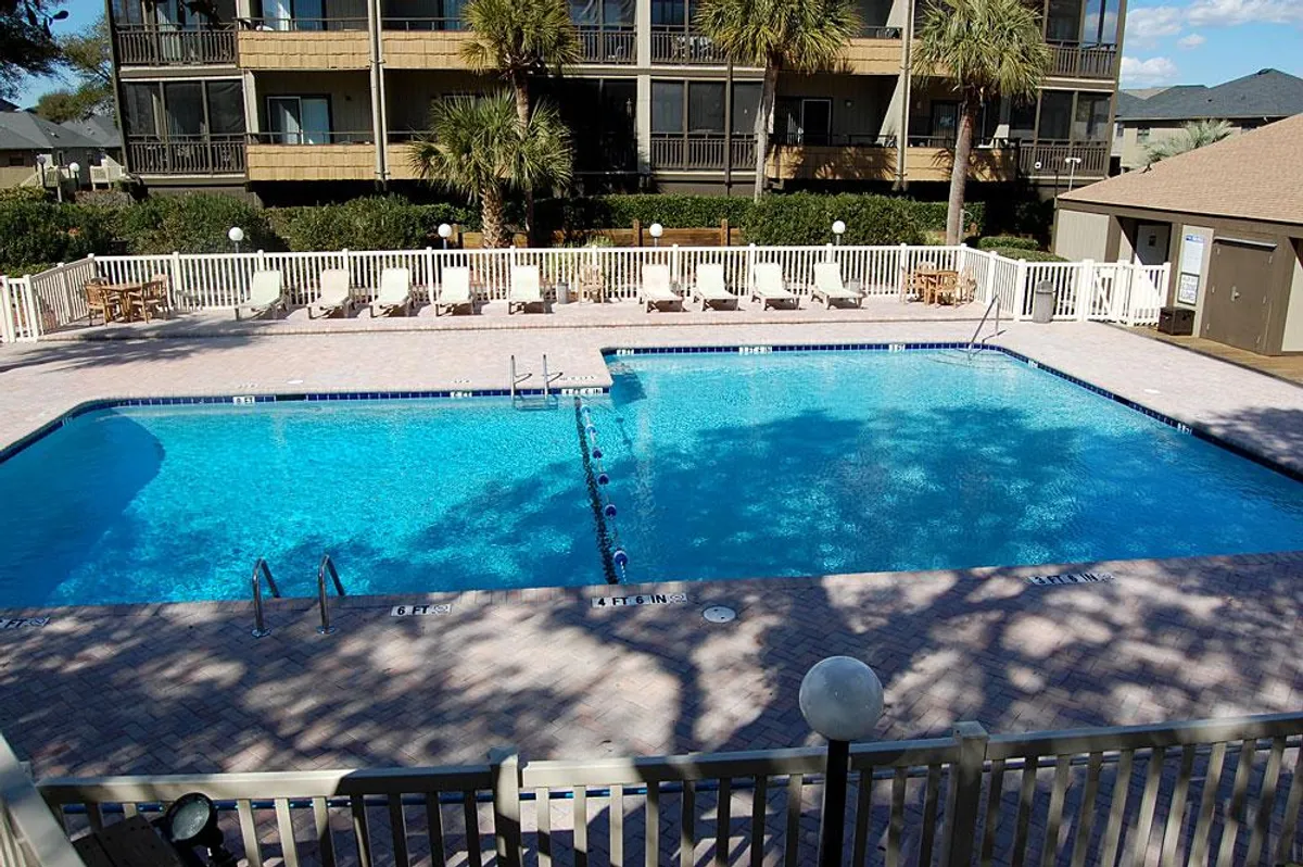 A lively outdoor swimming pool area surrounded by palm trees, with lounge chairs placed invitingly around the water. The pool sits adjacent to the Mariners Cove buildings, suggesting a fun, sun-soaked communal environment perfect for social gatherings and relaxation.
