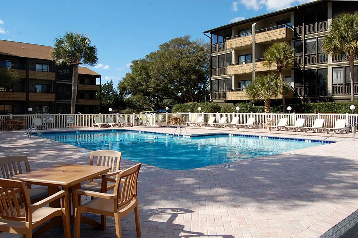 This image captures the Mariners Cove outdoor swimming pool surrounded by lounge chairs, a dining table set for two, and lush palm trees. Balconies of the buildings overlook the pool, suggesting a cozy atmosphere for couples to relax and connect outdoors.