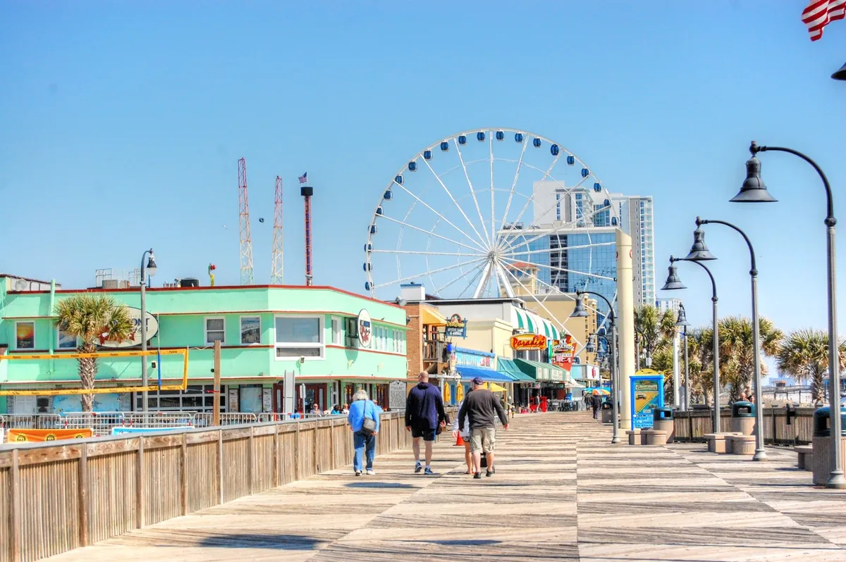 A vibrant scene from the Myrtle Beach boardwalk featuring the iconic ferris wheel, colorful buildings, and people strolling and engaging with the lively surroundings. The atmosphere exudes excitement and leisure, emphasizing the array of local attractions just a short drive away from Mariners Cove.