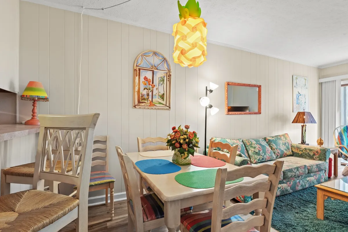 A colorful dining area highlighted by a playful pineapple light fixture, a cheerful dining table, and a floral couch nearby. The space feels open and inviting, with wall art and decor that encourage family meals and social gatherings.