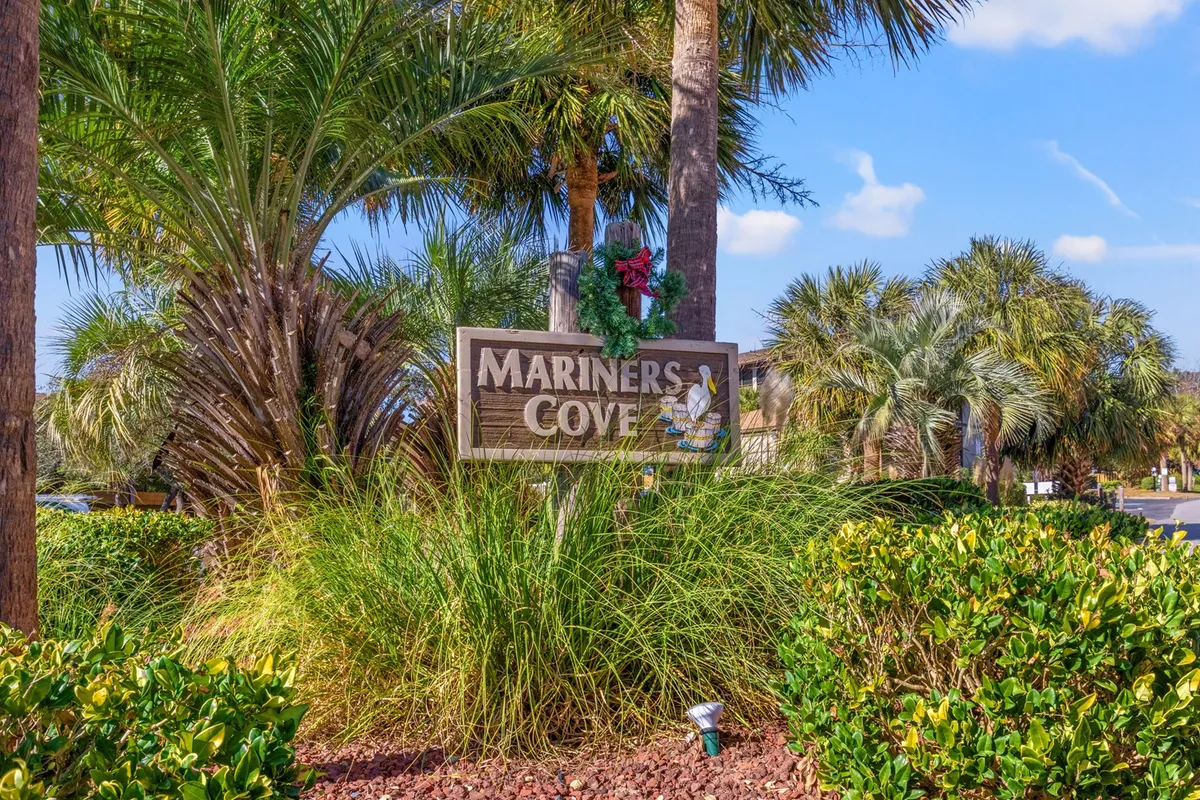 The Mariners Cove community entrance is marked by a welcoming sign surrounded by lush palm trees and green shrubs, suggesting a vibrant and friendly neighborhood setting.