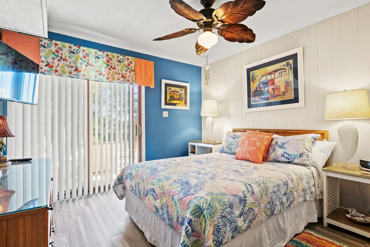 This image presents a bed with a colorful bedspread, flanked by matching lamps on bedside tables, a decorative ceiling fan overhead, and cheerful artwork on the wall. The space is clearly inviting and styled for comfort, radiating a cozy, restful ambiance ideal for a primary bedroom retreat.