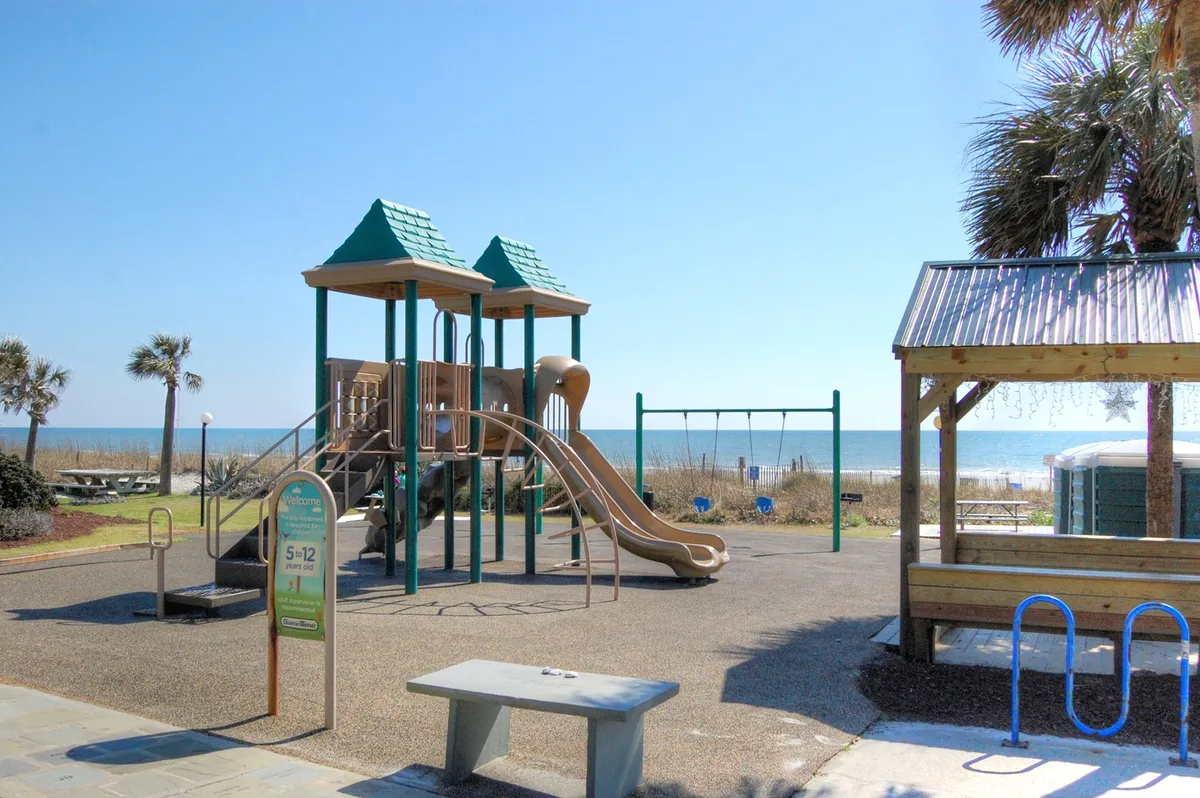A vibrant playground set against a picturesque beach backdrop, featuring swings, climbing equipment, a picnic area shaded by palm trees, and the sparkling ocean just steps away. Children are seen playing and families are relaxing, creating a lively, joyful atmosphere.